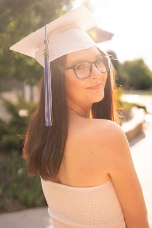 Graduate in white dress and cap smiles in warm sunlight during outdoor graduation photo session.