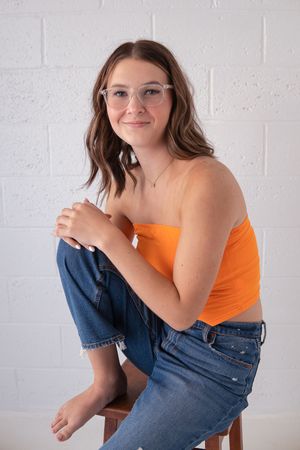 Casual portrait against white wall wearing orange top, jeans and glasses while sitting on wooden stool.