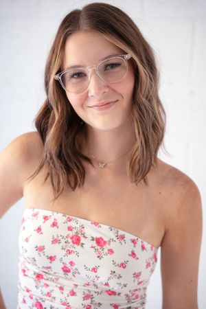 A person in a floral strapless top smiles warmly while wearing round glasses against a light background.