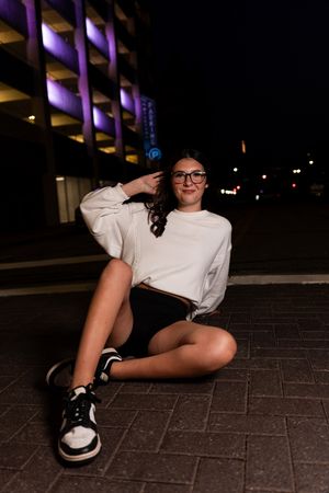 A person in a white t-shirt and black shoes lies on a dark brick surface at night under purple lighting.