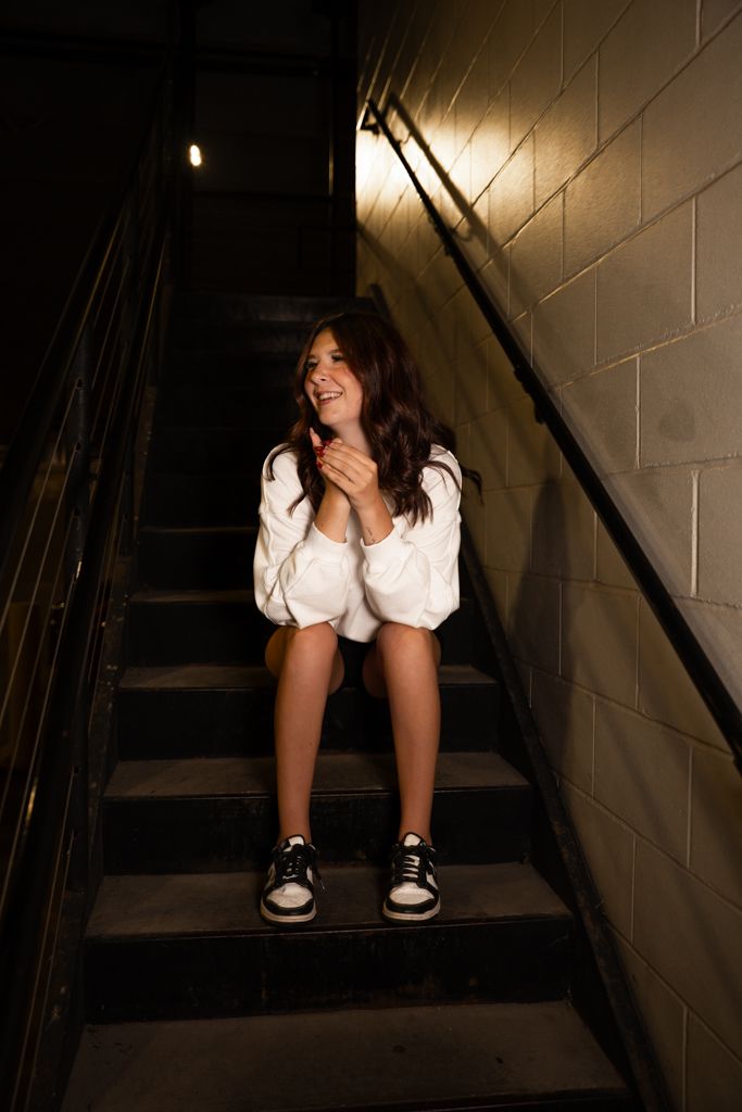 A person in an oversized white sweater and sneakers sits on dark concrete stairs in moody lighting.