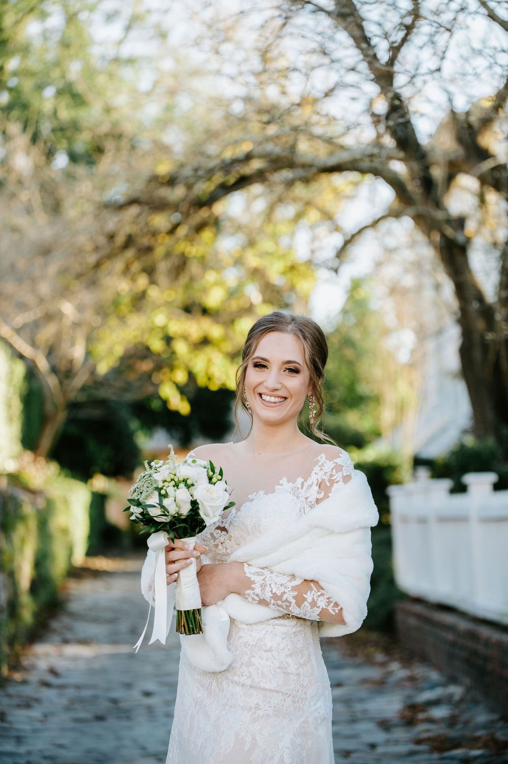 A bride in a white lace gown poses with a bouquet in a garden setting during an outdoor wedding photoshoot.