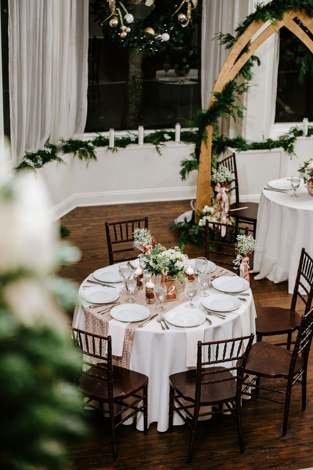 Elegant wedding reception tables with white linens, dark wooden chairs, and delicate floral centerpieces in a rustic venue.