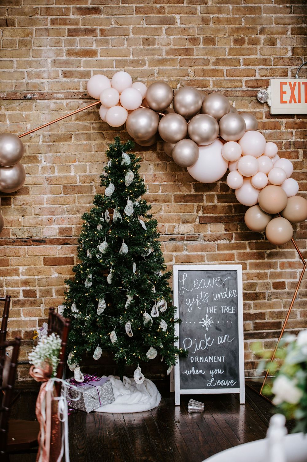 A Christmas tree decorated with silver ornaments under a pink and silver balloon arch against a brick wall.