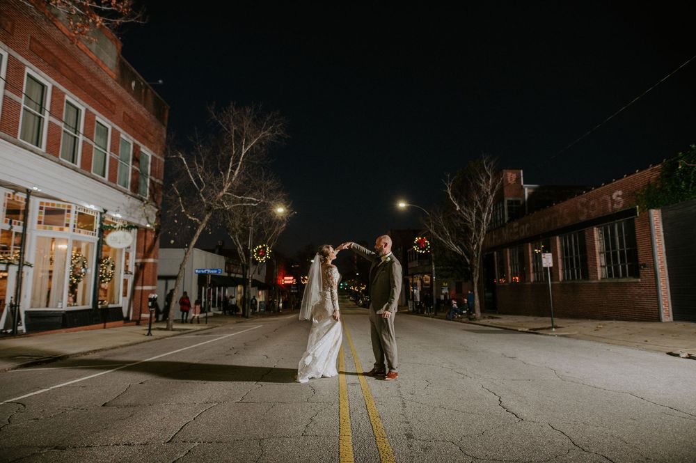 A couple in wedding attire stands in the middle of an empty street at night with illuminated storefronts in the background.