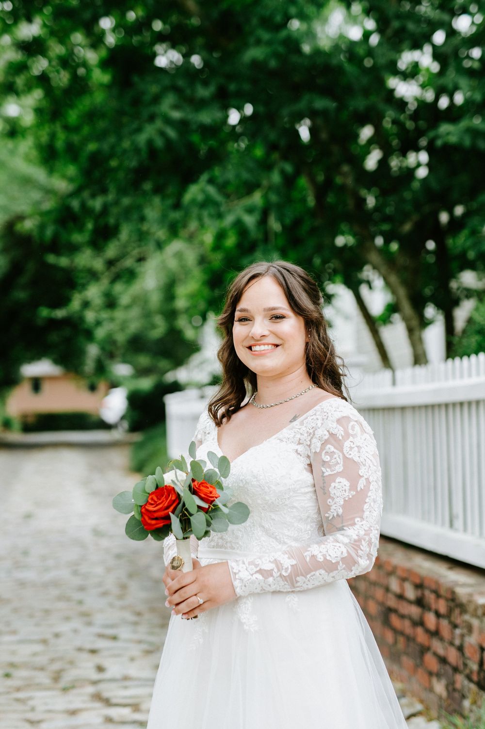 Bride in elegant lace sleeve wedding dress poses outdoors near white picket fence and greenery with red rose bouquet.