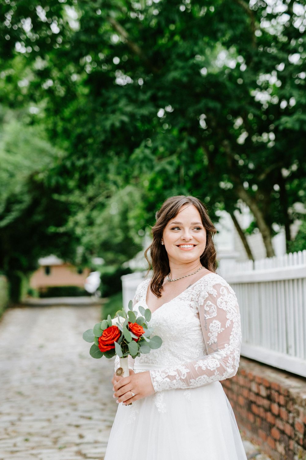 A bride in a white lace wedding dress with long sleeves holds red roses against a white picket fence background.