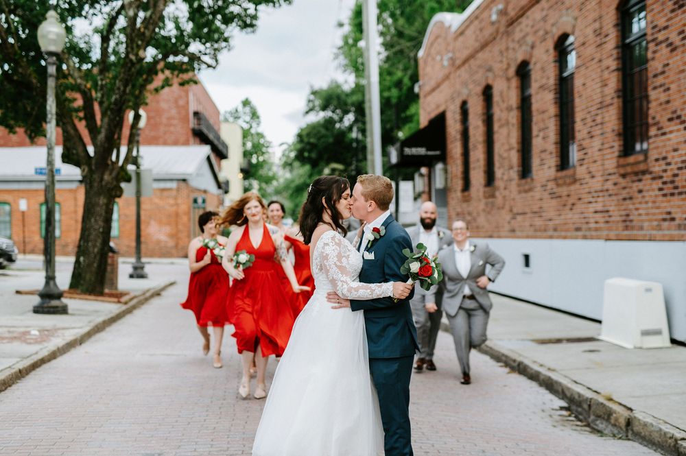 Wedding party in red dresses and formal attire walk down a historic brick street on a sunny day.