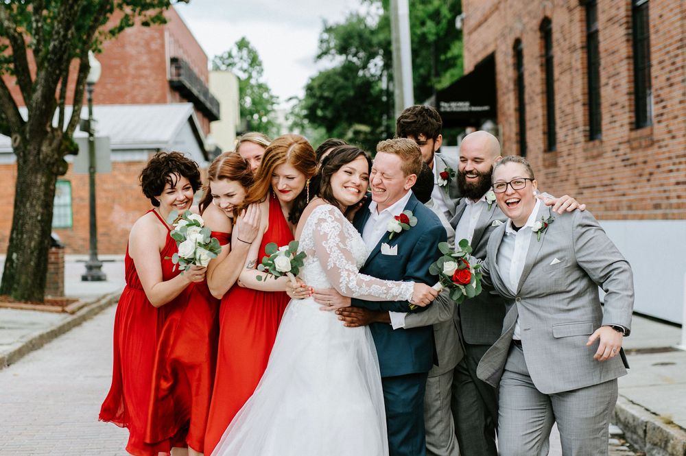 Wedding party in red bridesmaid dresses and gray suits pose playfully on a brick-lined street.