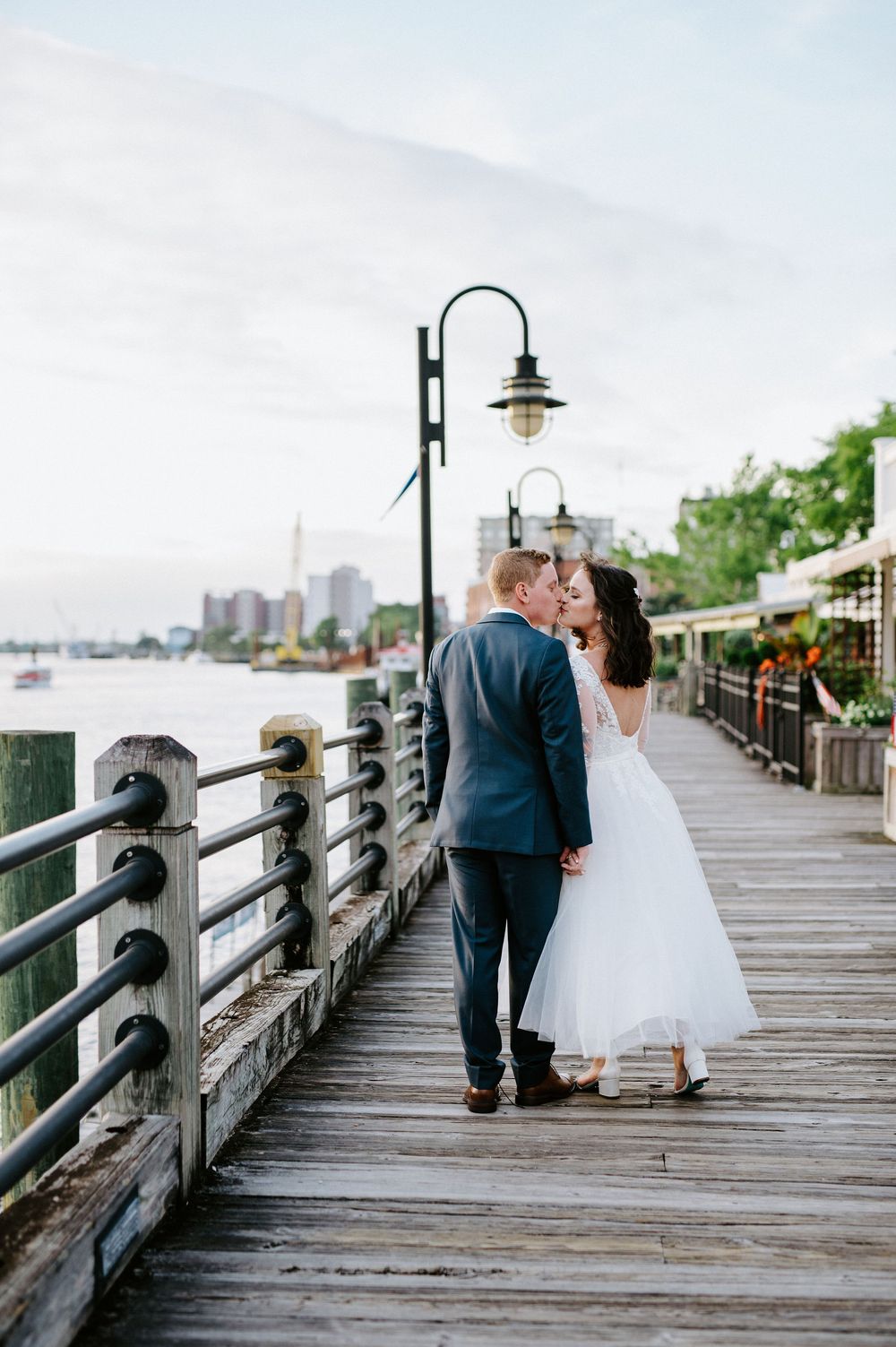 A couple in wedding attire walking along a wooden boardwalk with waterfront views.