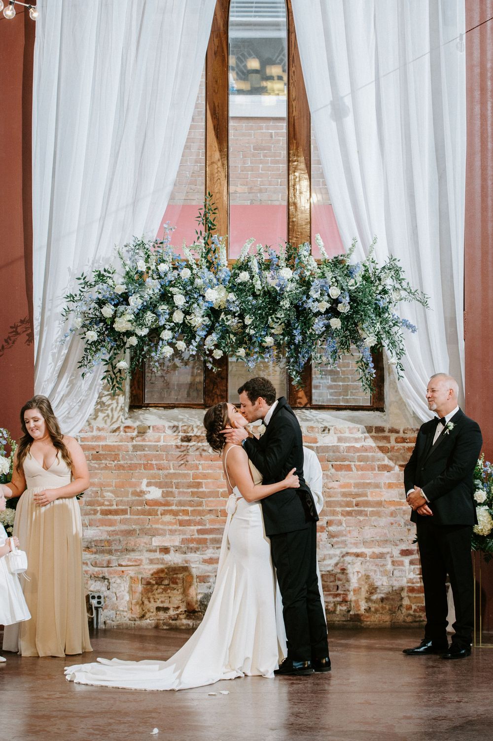 Wedding party stands in line during ceremony in elegant brick-walled venue with white floral decorations and drapes.