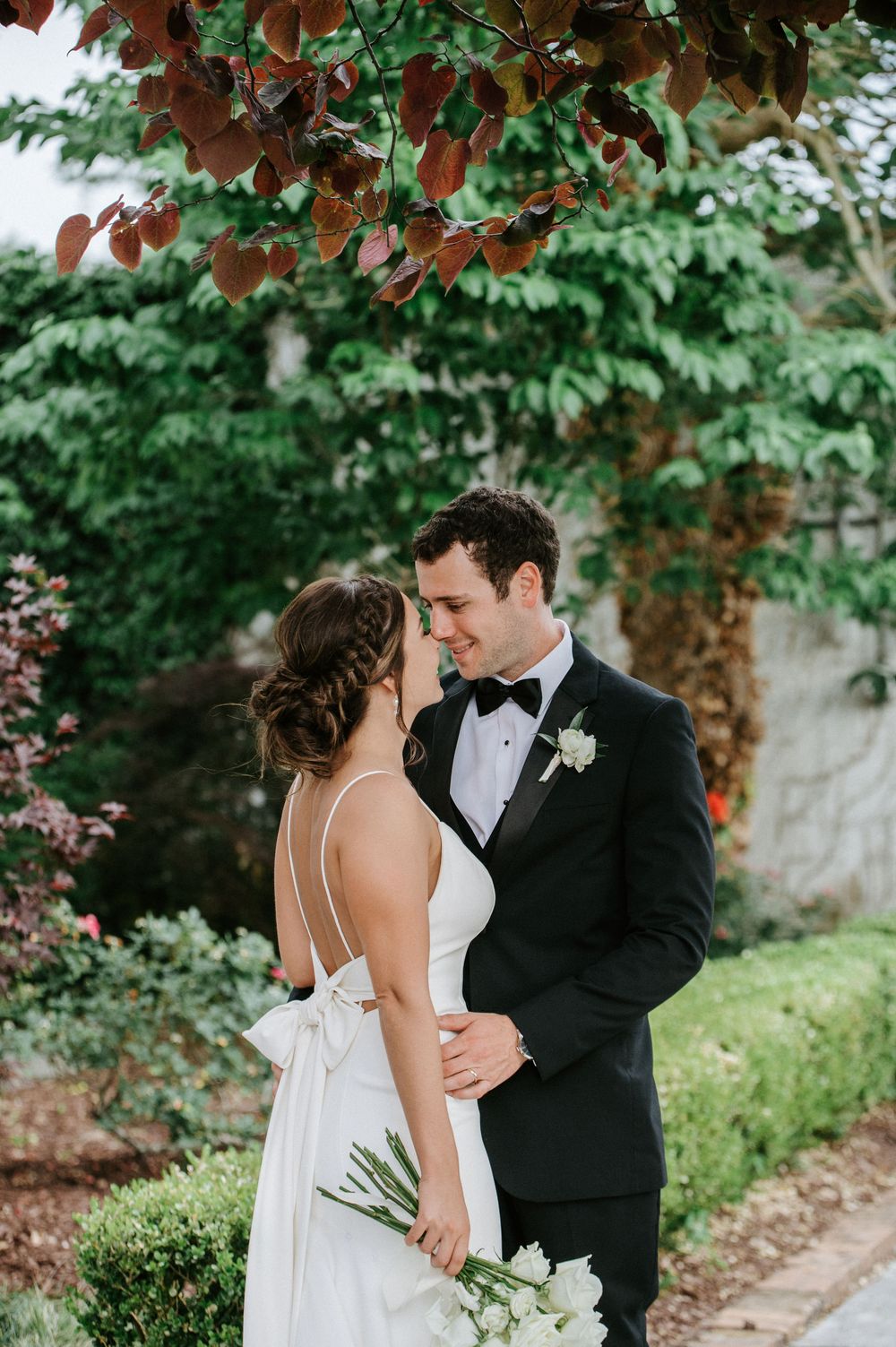 A romantic wedding moment captured in a lush garden setting with green foliage and autumn leaves overhead.