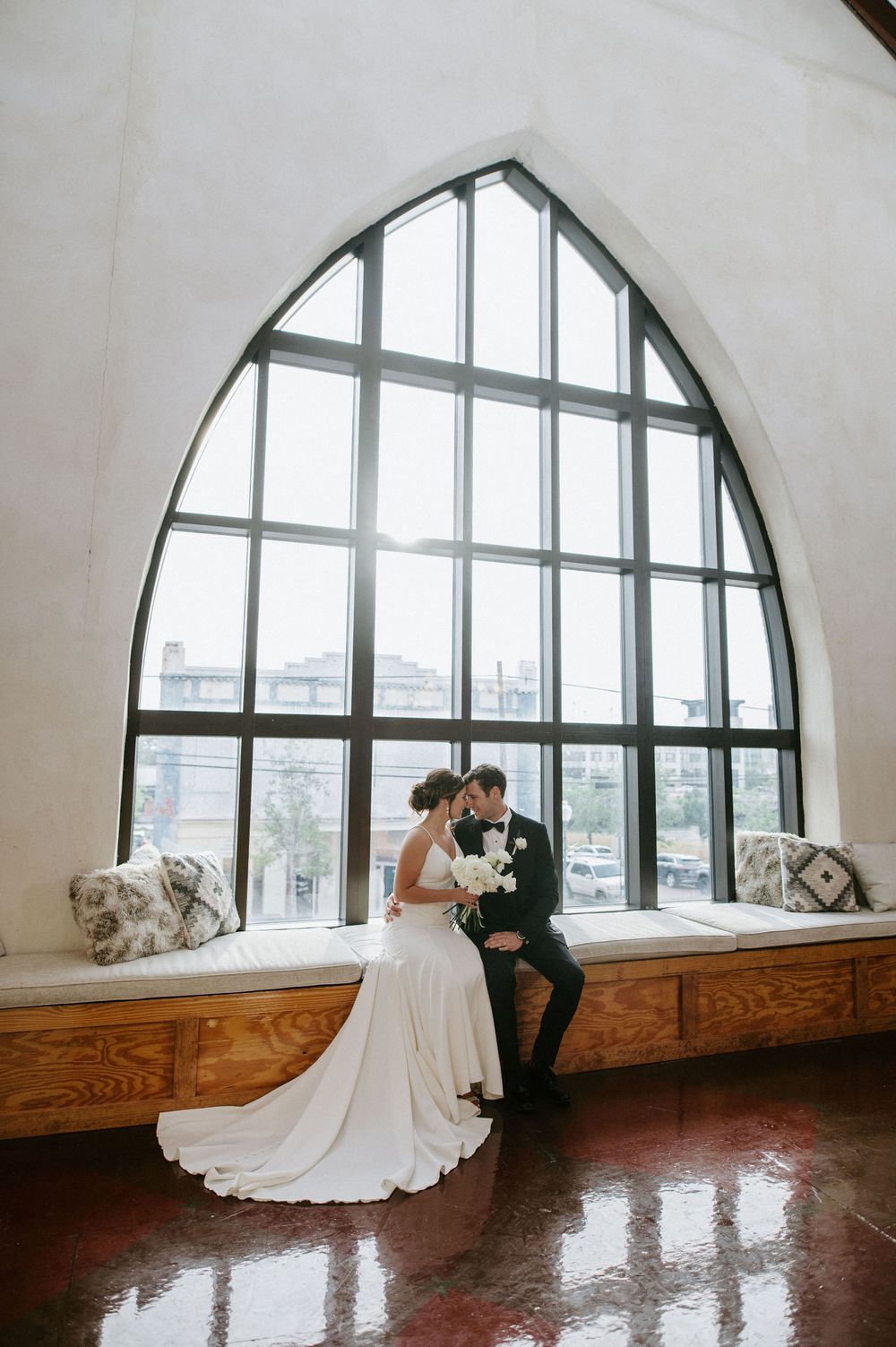 A wedding couple shares an intimate moment by a large arched window in an elegant venue.