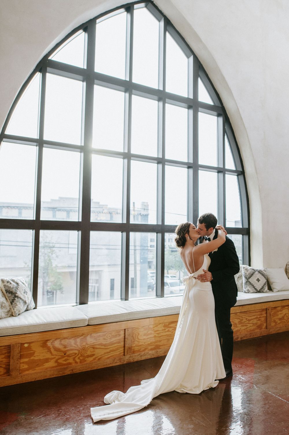 A romantic wedding scene in front of a grand arched window with natural light streaming in and wooden bench seating.