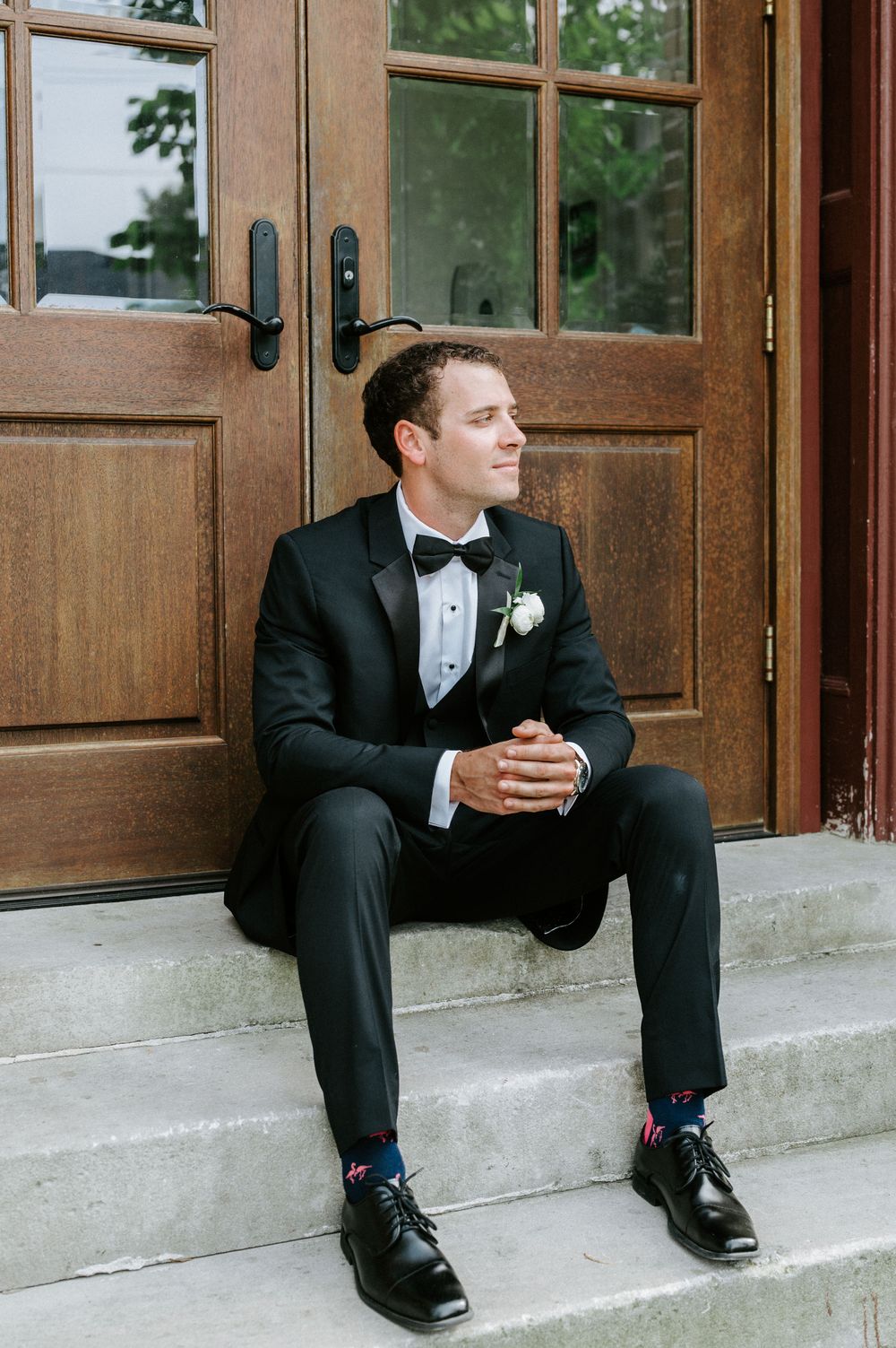 A groom in a black tuxedo and bow tie sits on stone steps near wooden doors, looking contemplatively to the side.