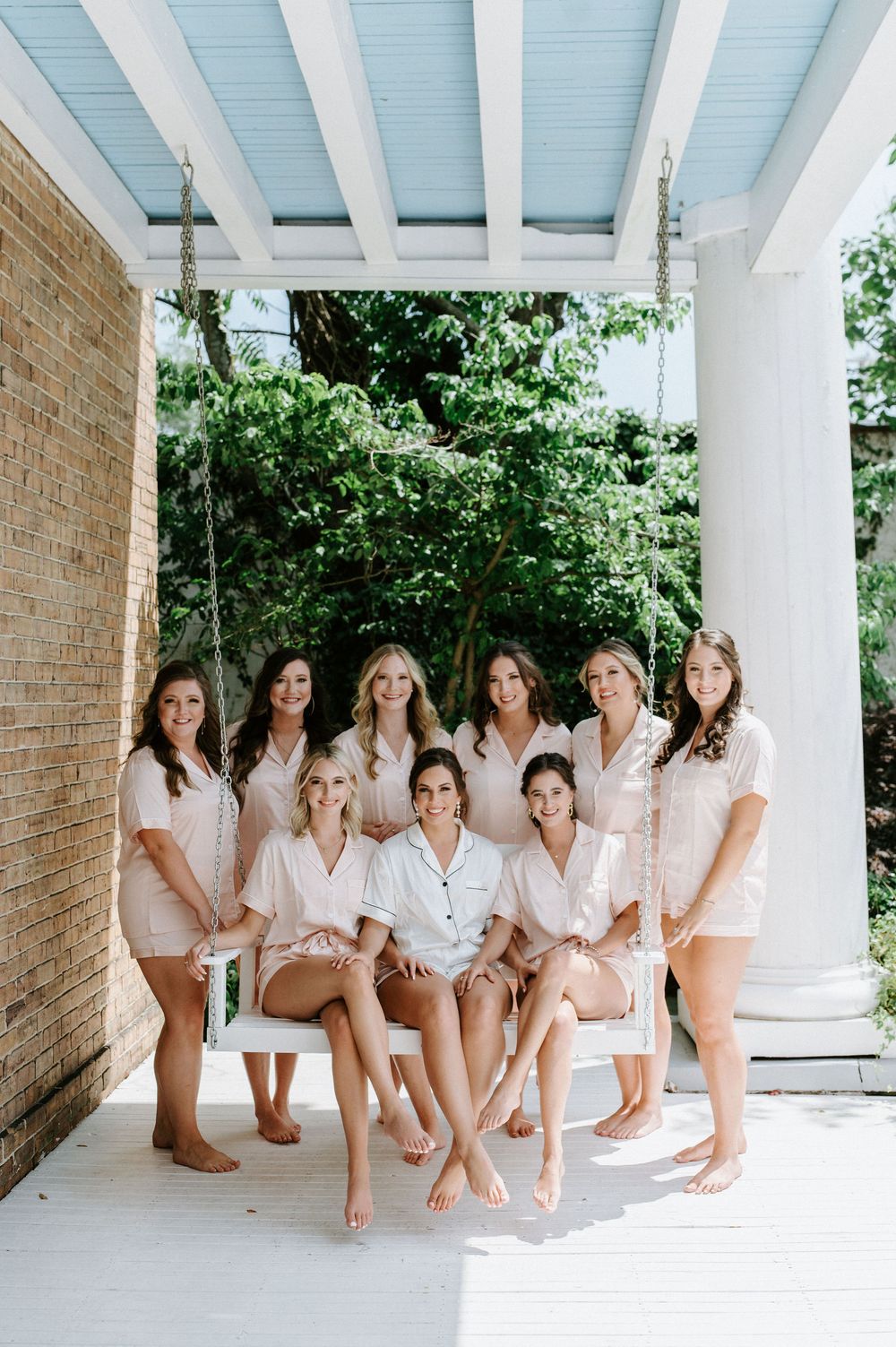A group in matching white robes sits together on a covered outdoor porch with white columns and brick walls.