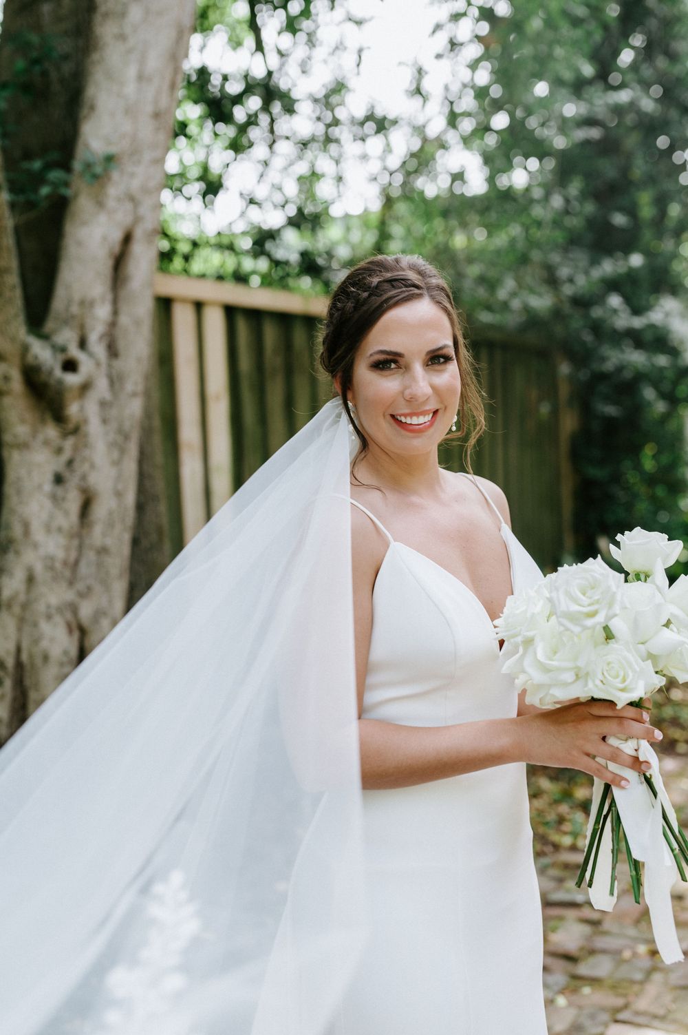 A bride in a white wedding dress holds a bouquet of white roses in an outdoor garden setting.