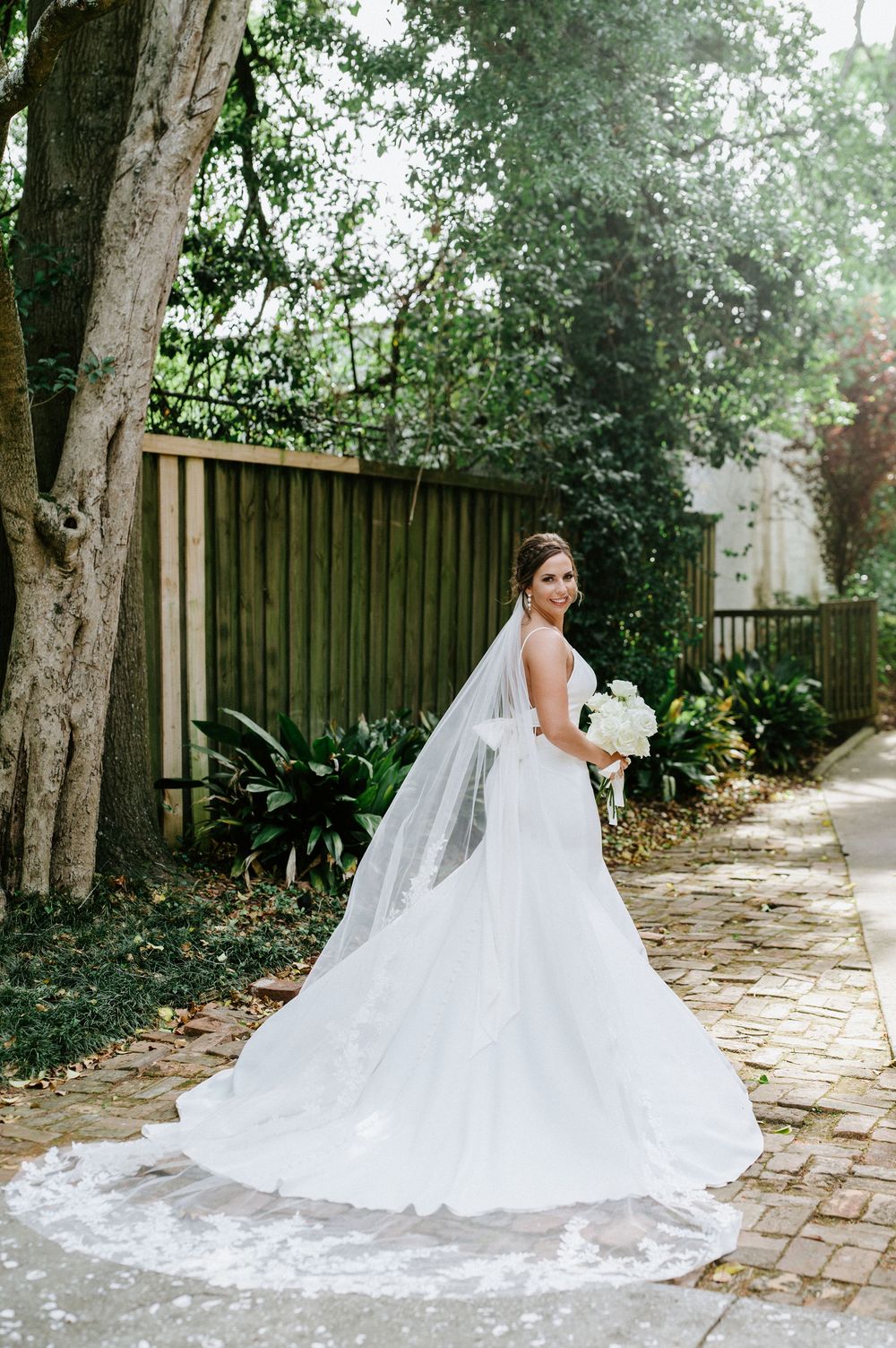 A bride in a flowing white wedding gown and long veil poses gracefully on a brick pathway surrounded by trees.