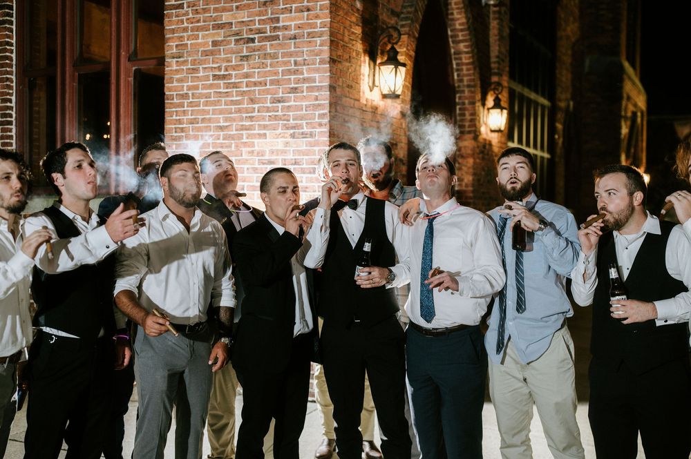 Groomsmen in suits and vests celebrate with raised drinks during a wedding reception in a rustic brick venue.