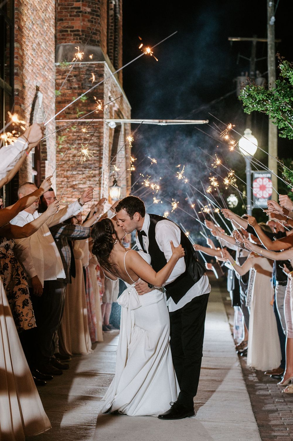 Wedding party lines up holding sparklers creating a magical light tunnel effect against brick walls.