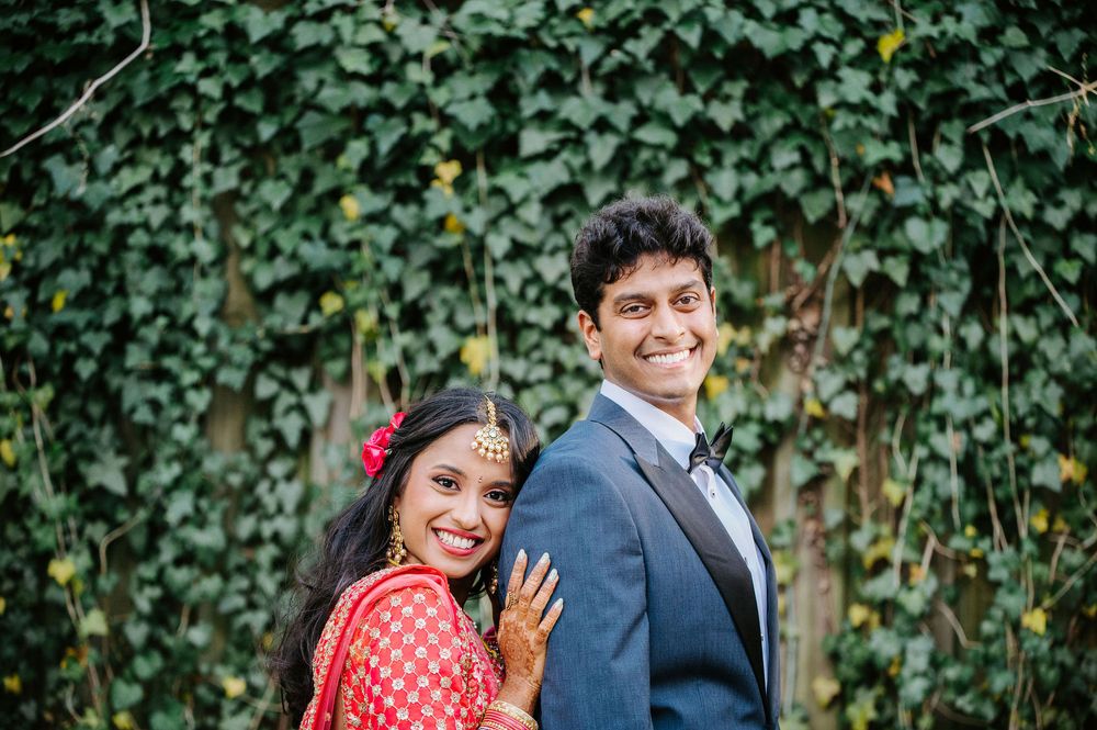 A couple in traditional Indian wedding attire share a joyful moment against a green ivy backdrop.