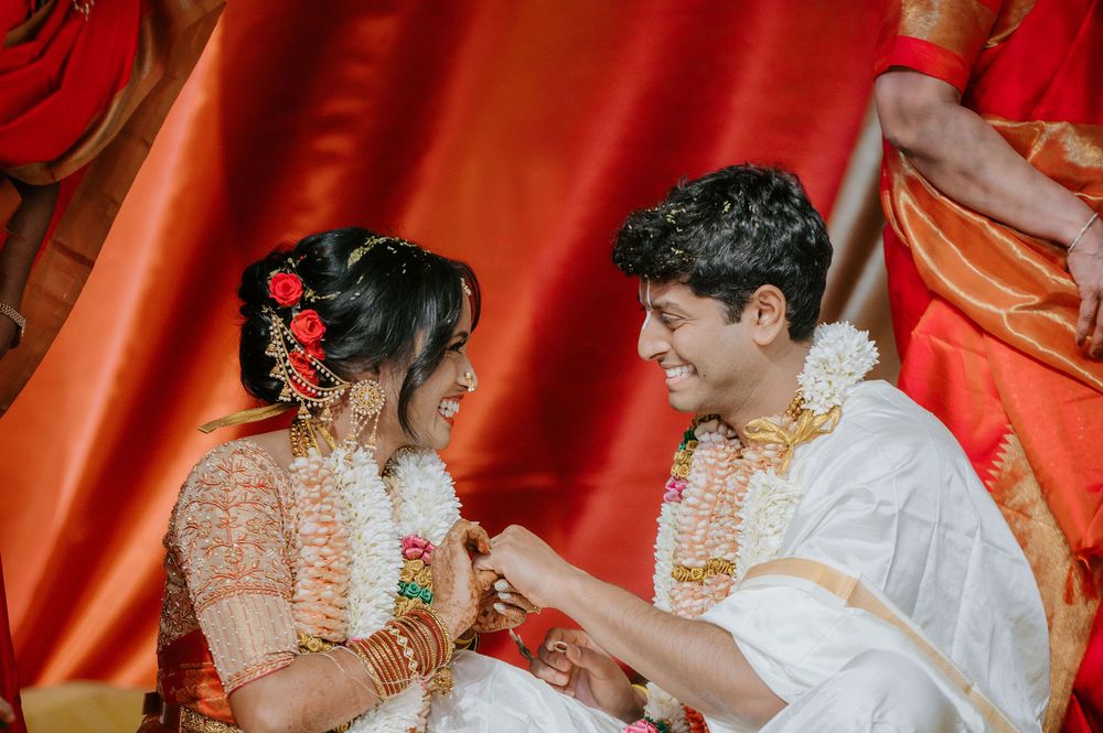 A couple in traditional Indian wedding attire performs ceremonial rituals with red and gold decor in the background.