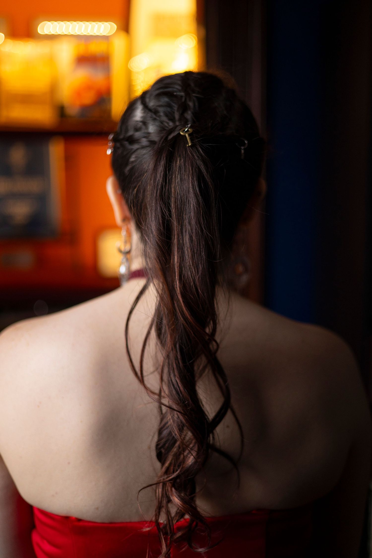 Back view of a bride with long dark curled hair styled in a ponytail against a red strapless dress with warm ambient lighting.