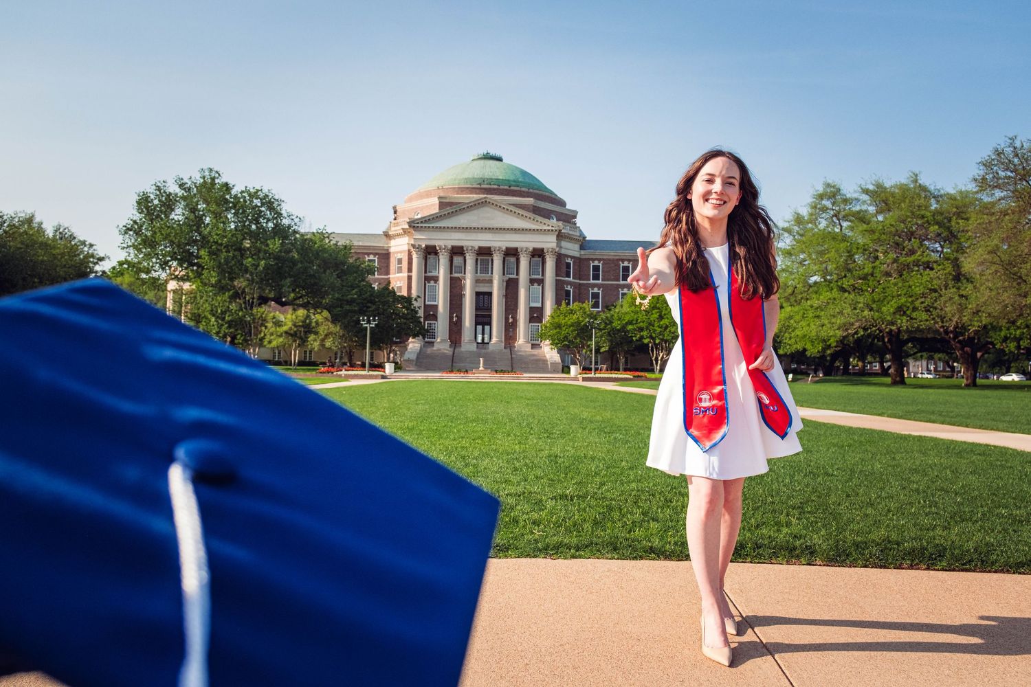 Graduate in white dress and red stole poses joyfully in front of a domed university building.