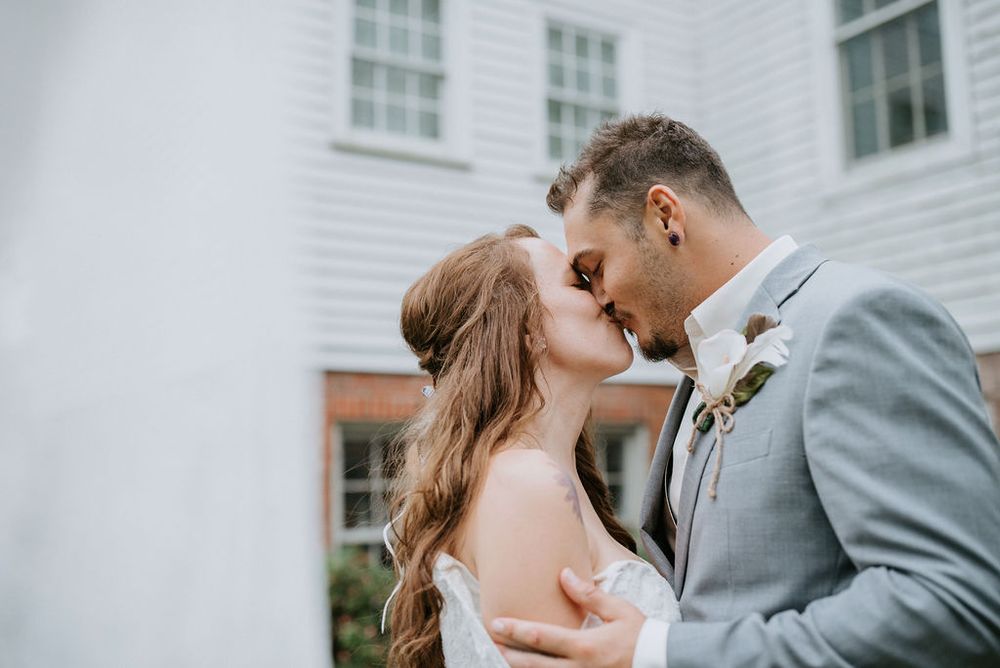A couple shares a romantic kiss on their wedding day with a white building in the background.