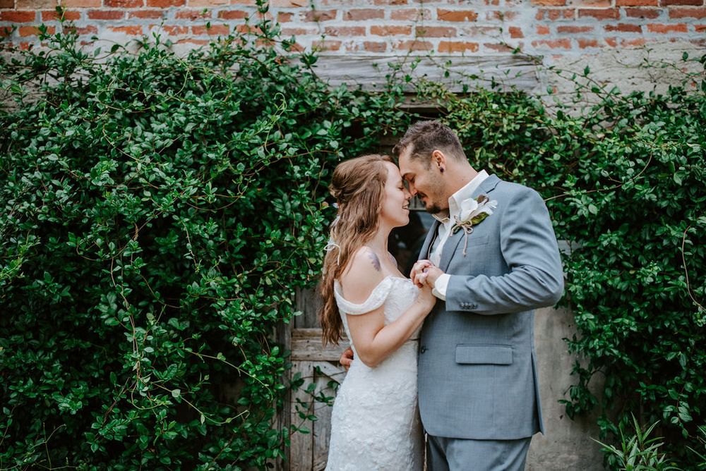 Romantic wedding couple embraces in front of a rustic brick wall with lush green foliage.
