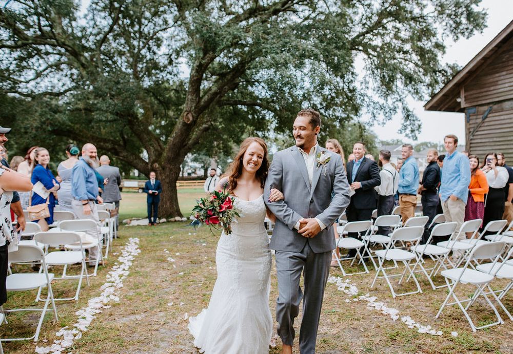 Couple walking down an aisle lined with white chairs under oak trees at an outdoor wedding ceremony.