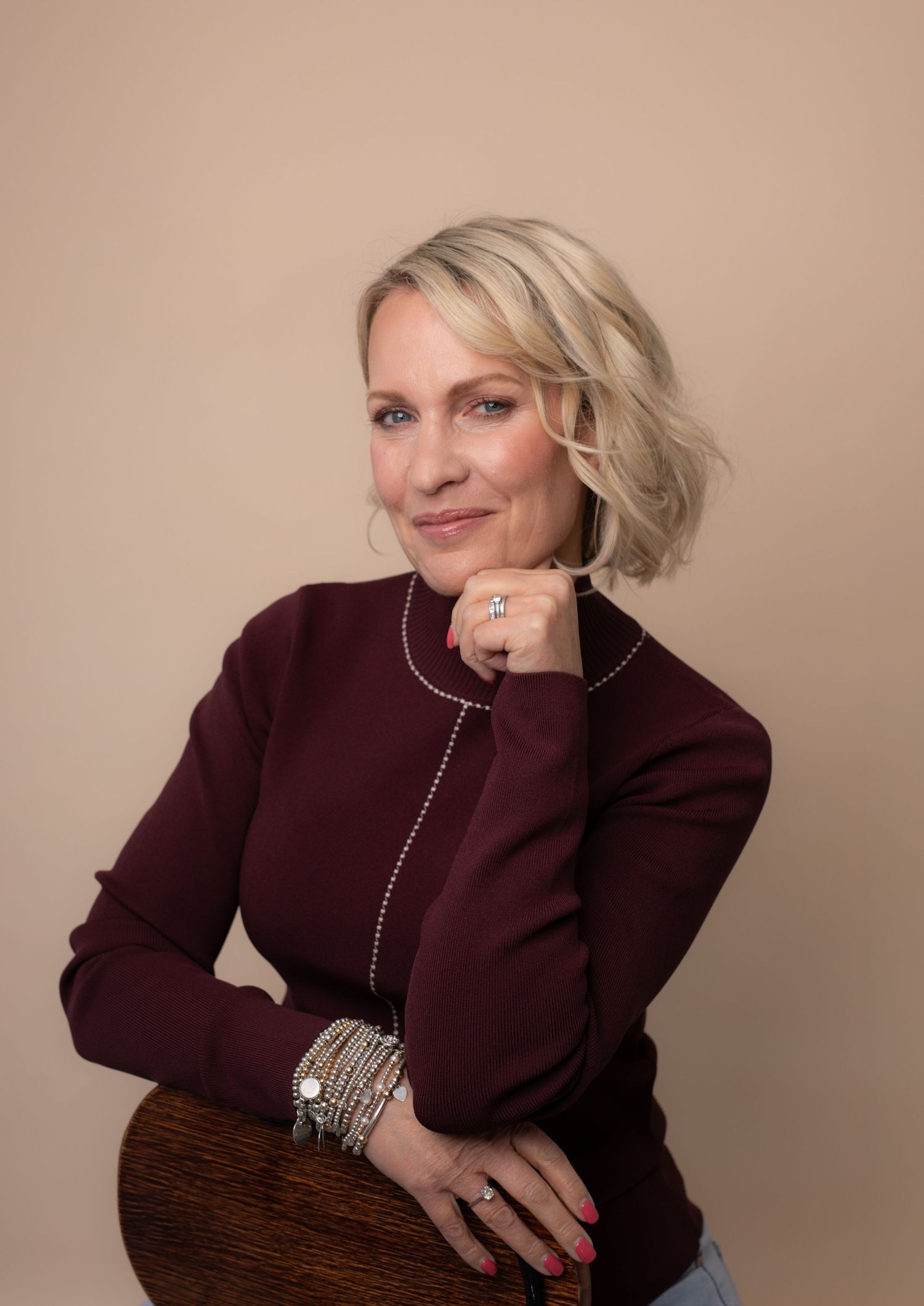 Blonde woman in burgundy turtleneck sweater poses confidently with hand on chin, wearing silver bracelets and rings.