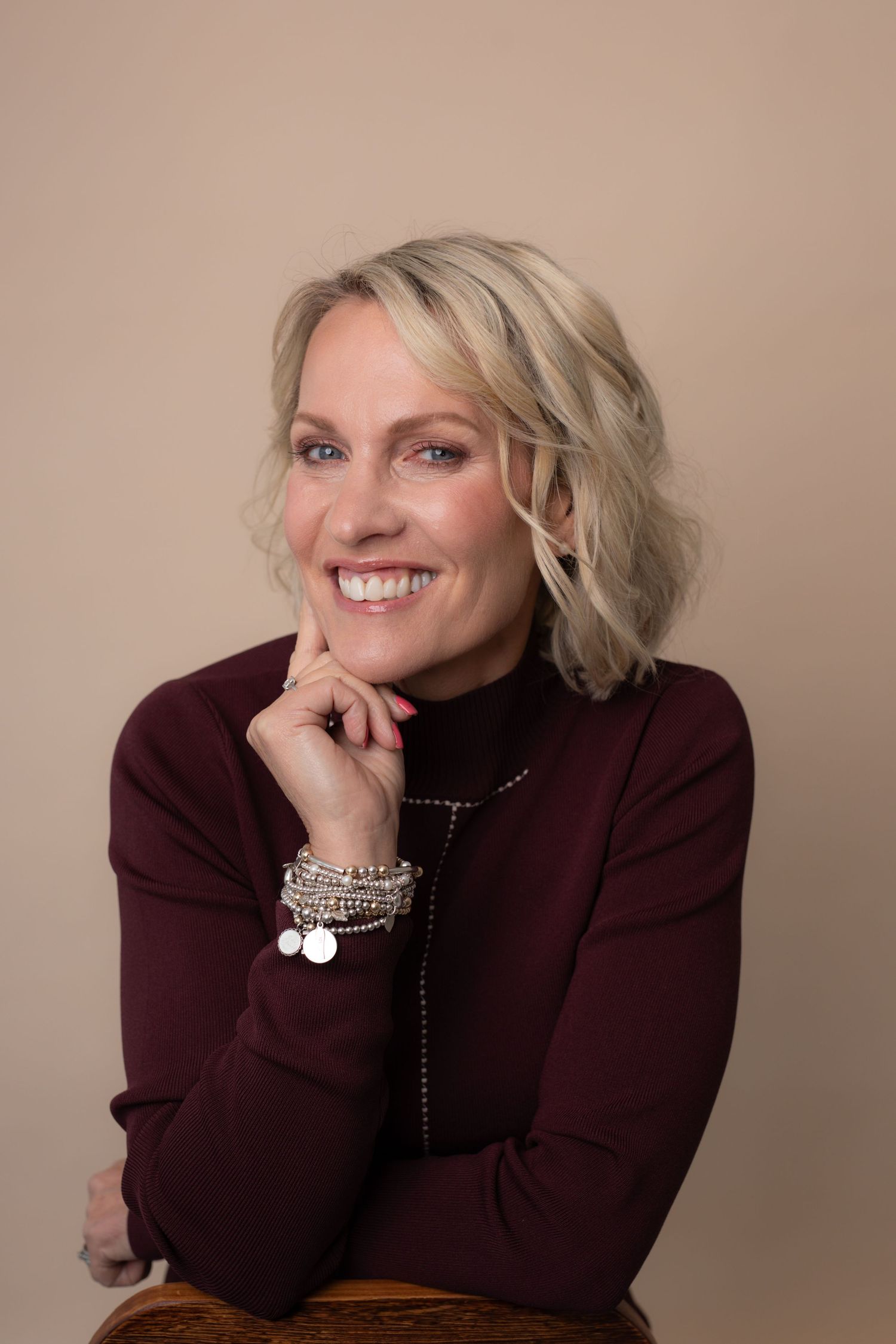 Smiling blonde woman in burgundy turtleneck with layered bracelets poses confidently against a neutral beige background.