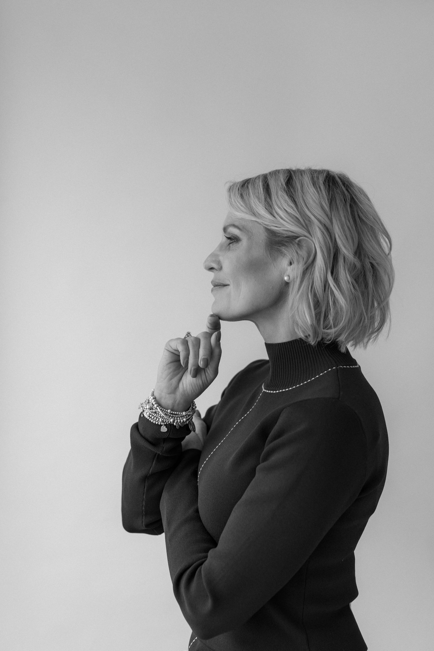 Black and white profile portrait of a thoughtful blonde woman in a turtleneck, hand on chin, wearing bracelets and earrings.