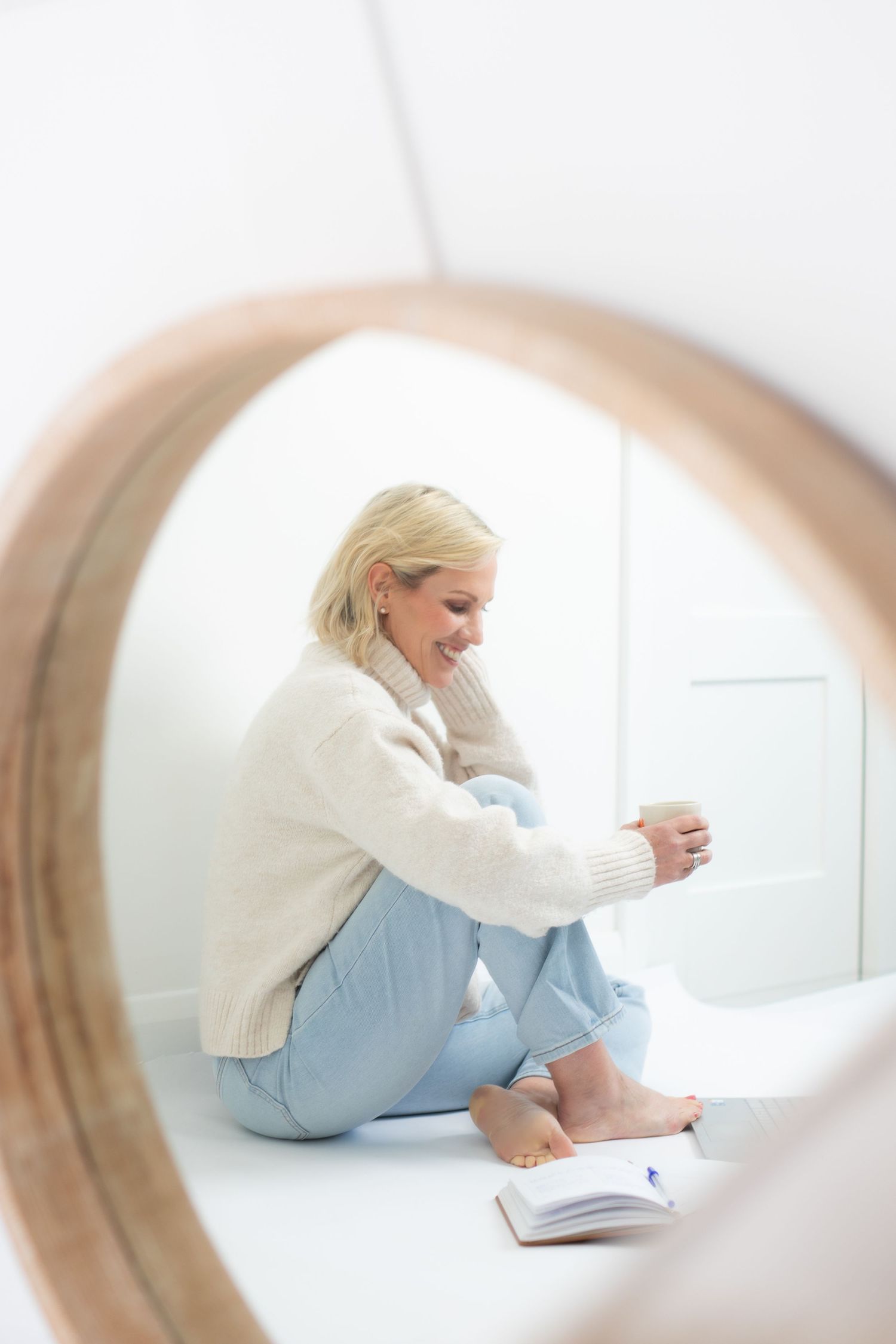 Smiling blonde woman in white sweater and jeans sitting on floor with book and coffee cup, viewed through wooden circle frame.