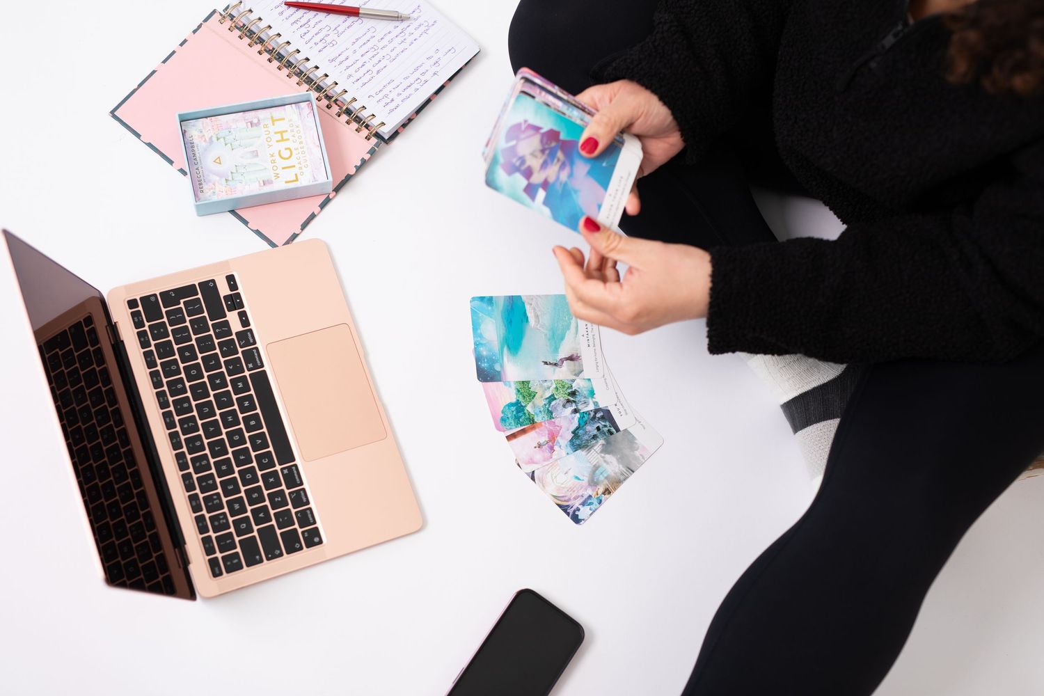 Woman sitting on white floor with laptop, phone, notebooks, and colorful art cards spread out around her.