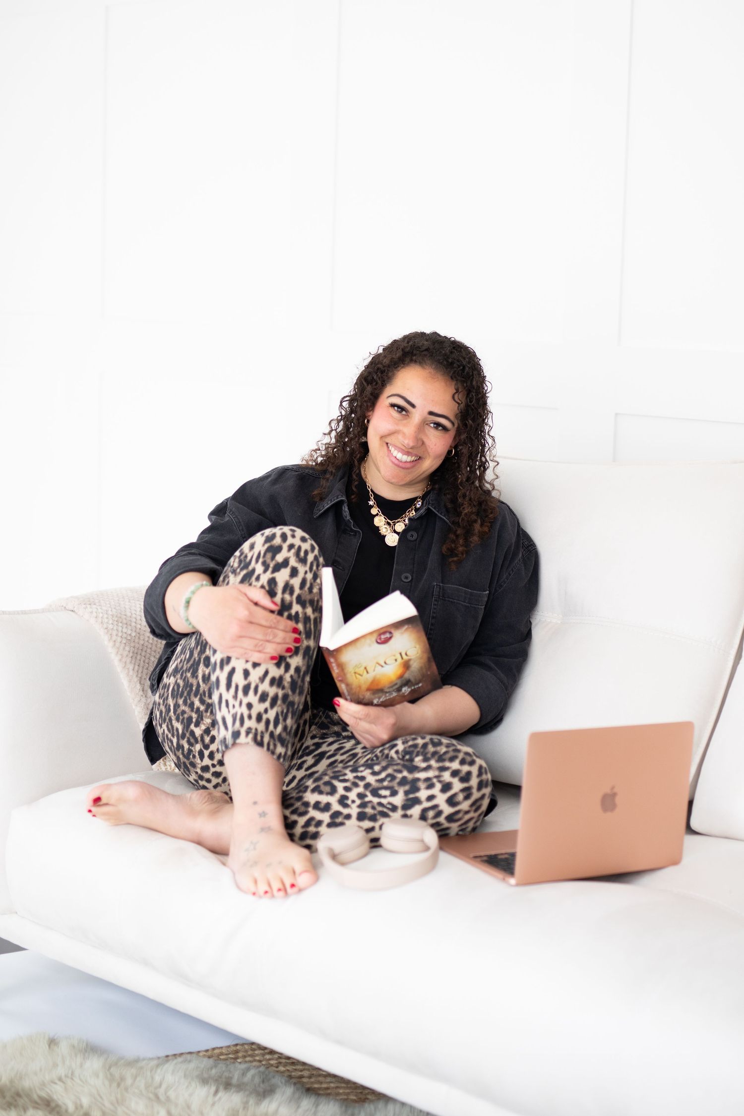 Woman smiling on white sofa with book and rose gold laptop, wearing leopard print pants and black jacket.