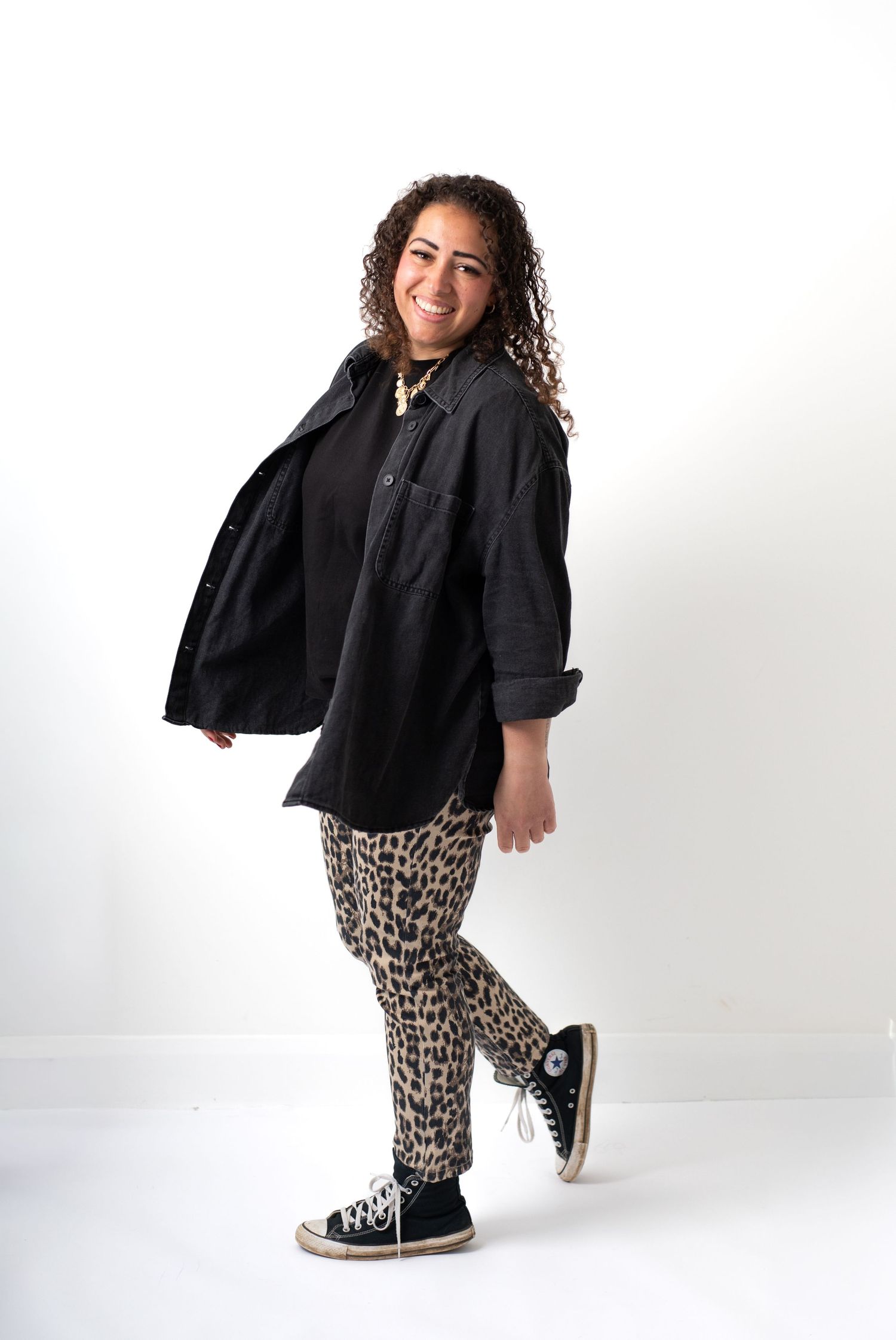 Woman smiling in black oversized shirt, leopard print pants, and black Converse sneakers on white background.