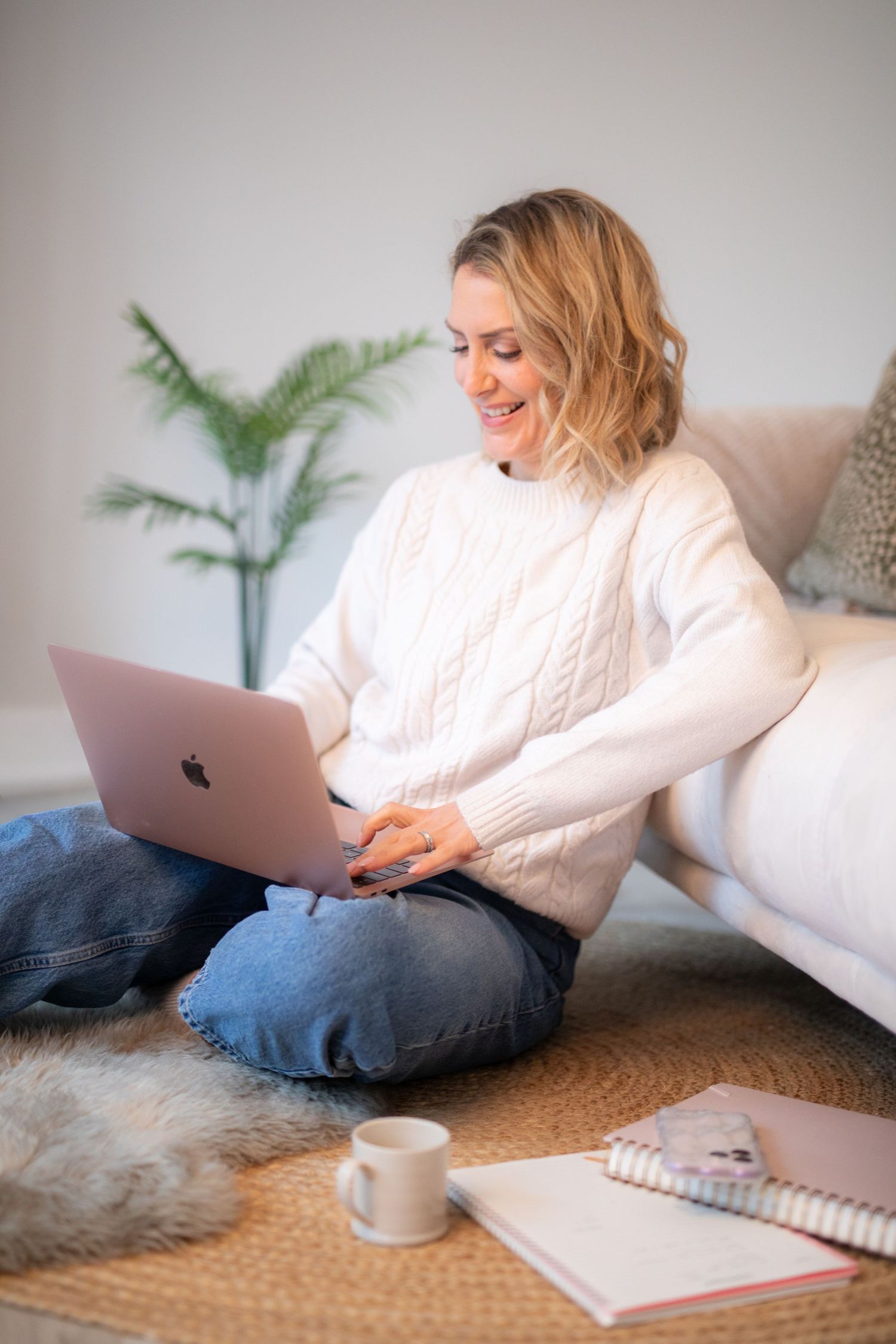 Person in cozy white sweater working on laptop while relaxing on floor with coffee and notebook nearby.
