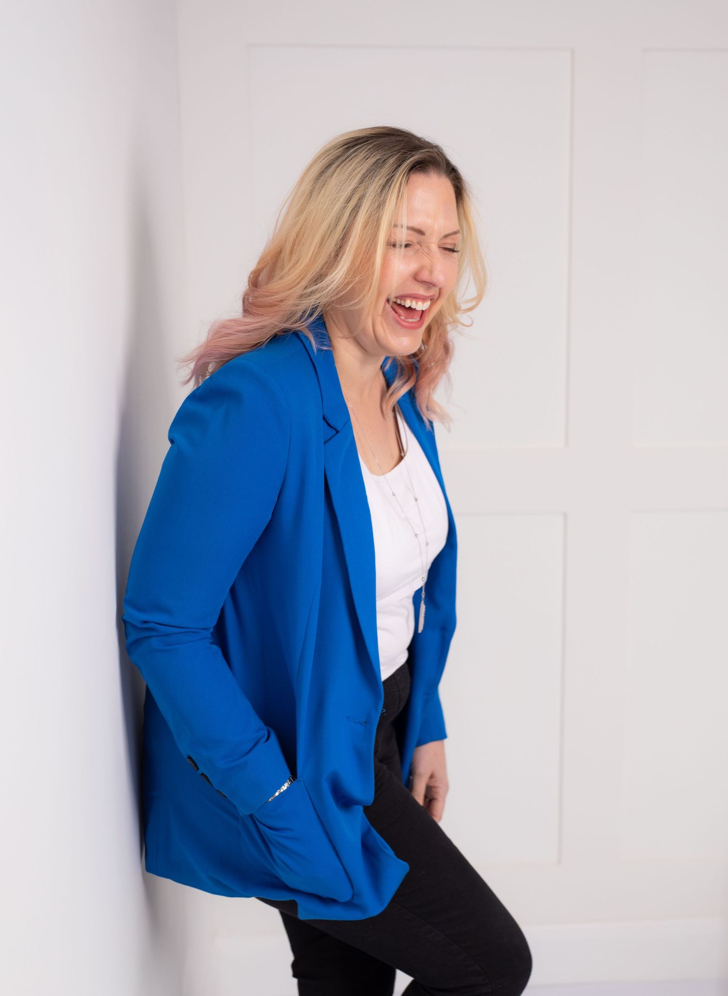 Professional wearing a vibrant royal blue blazer and white top laughs joyfully against a light background.
