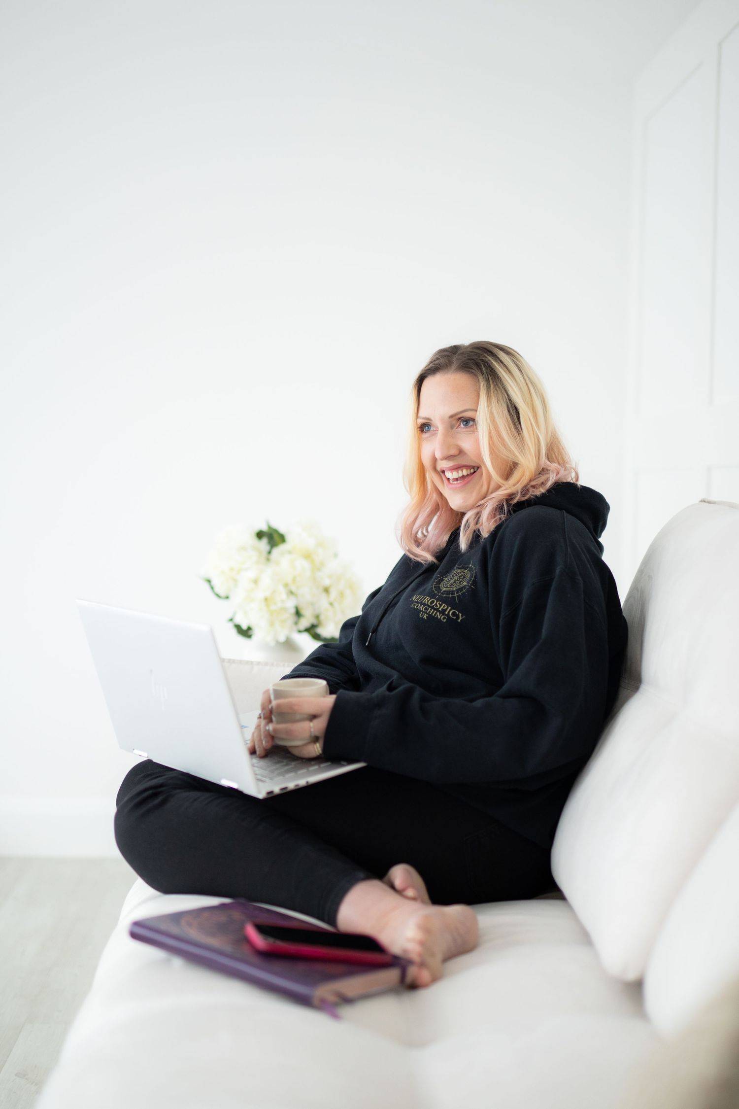Person in black loungewear relaxes on white couch with laptop and coffee in bright minimalist setting.