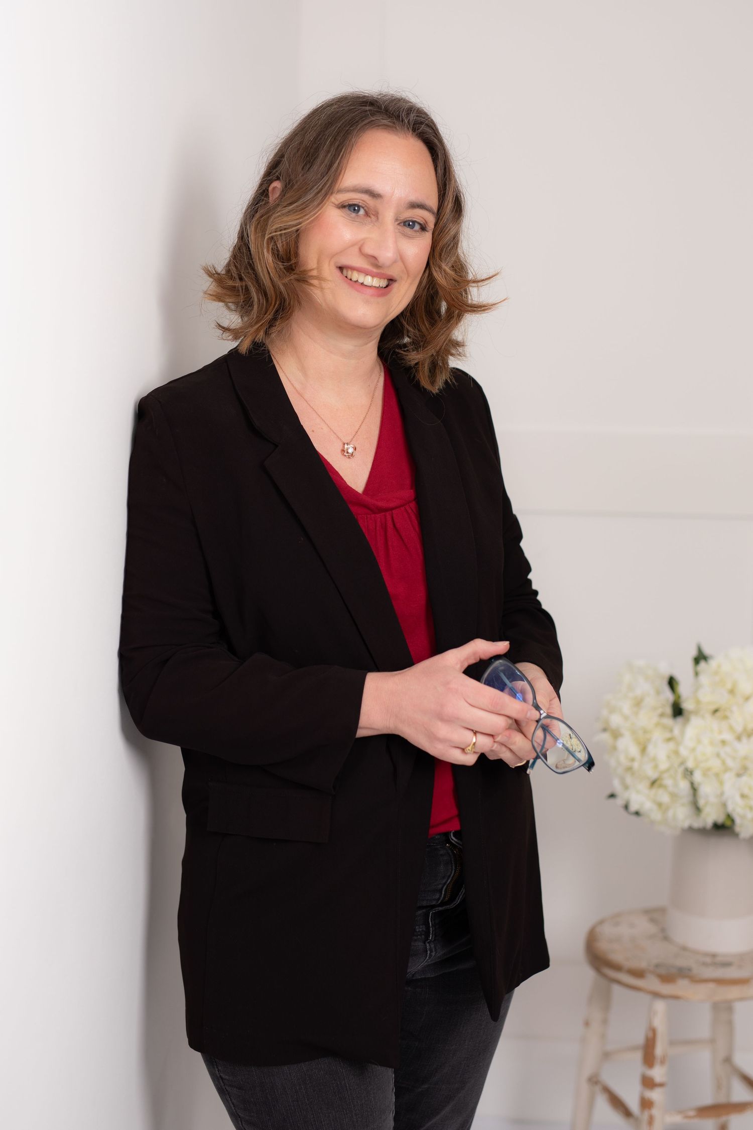 Professional woman in black blazer and red top poses in bright white studio setting with elegant decor.