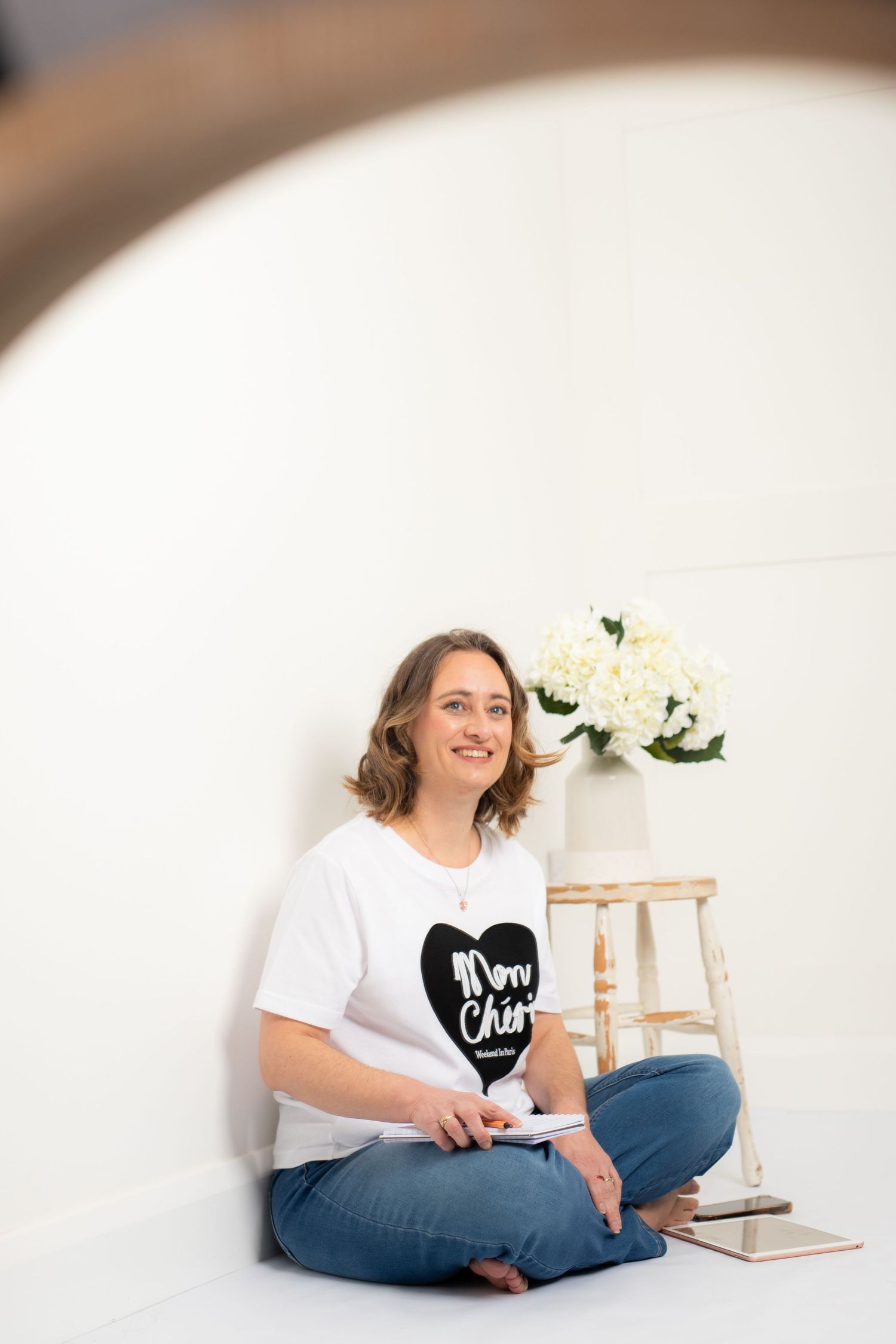 Woman in white t-shirt and jeans sits casually on floor with white flowers in modern minimalist setting.