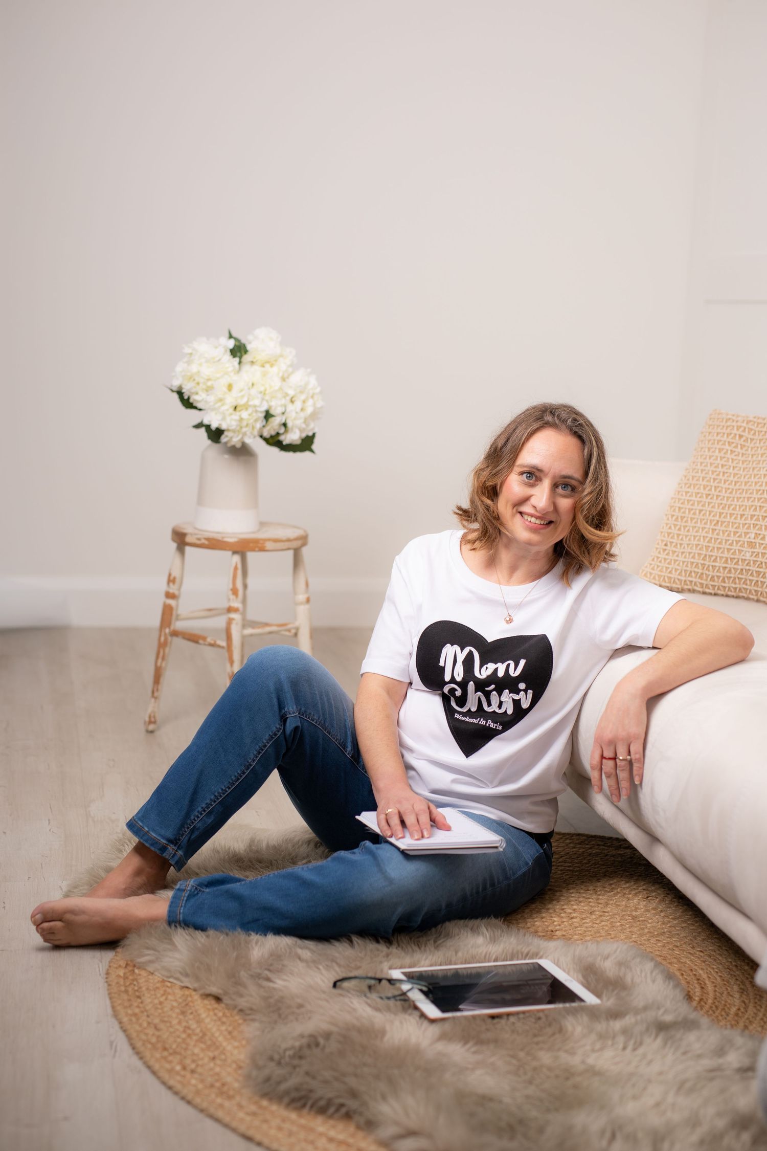 A relaxed person in jeans and a graphic t-shirt sits on the floor near a white couch with flowers on a wooden stool nearby.