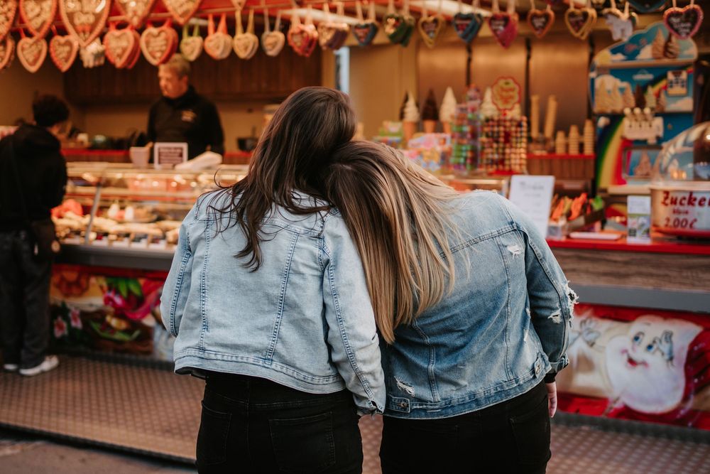 Two people in denim jackets stand close together at a holiday market with festive decorations and lights.