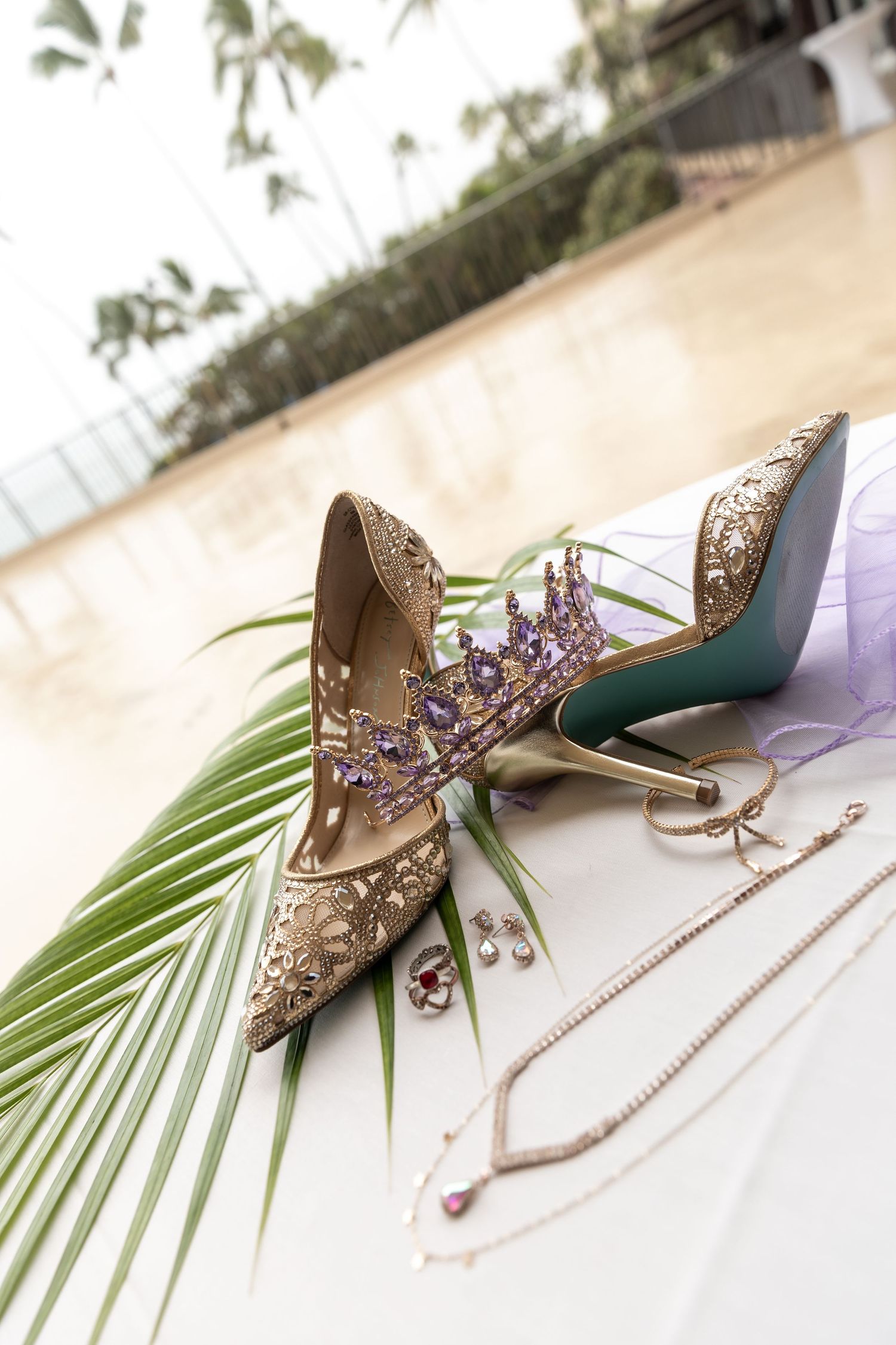 Elegant bridal shoes with crystal embellishments and lavender accents displayed on palm leaves against white background.