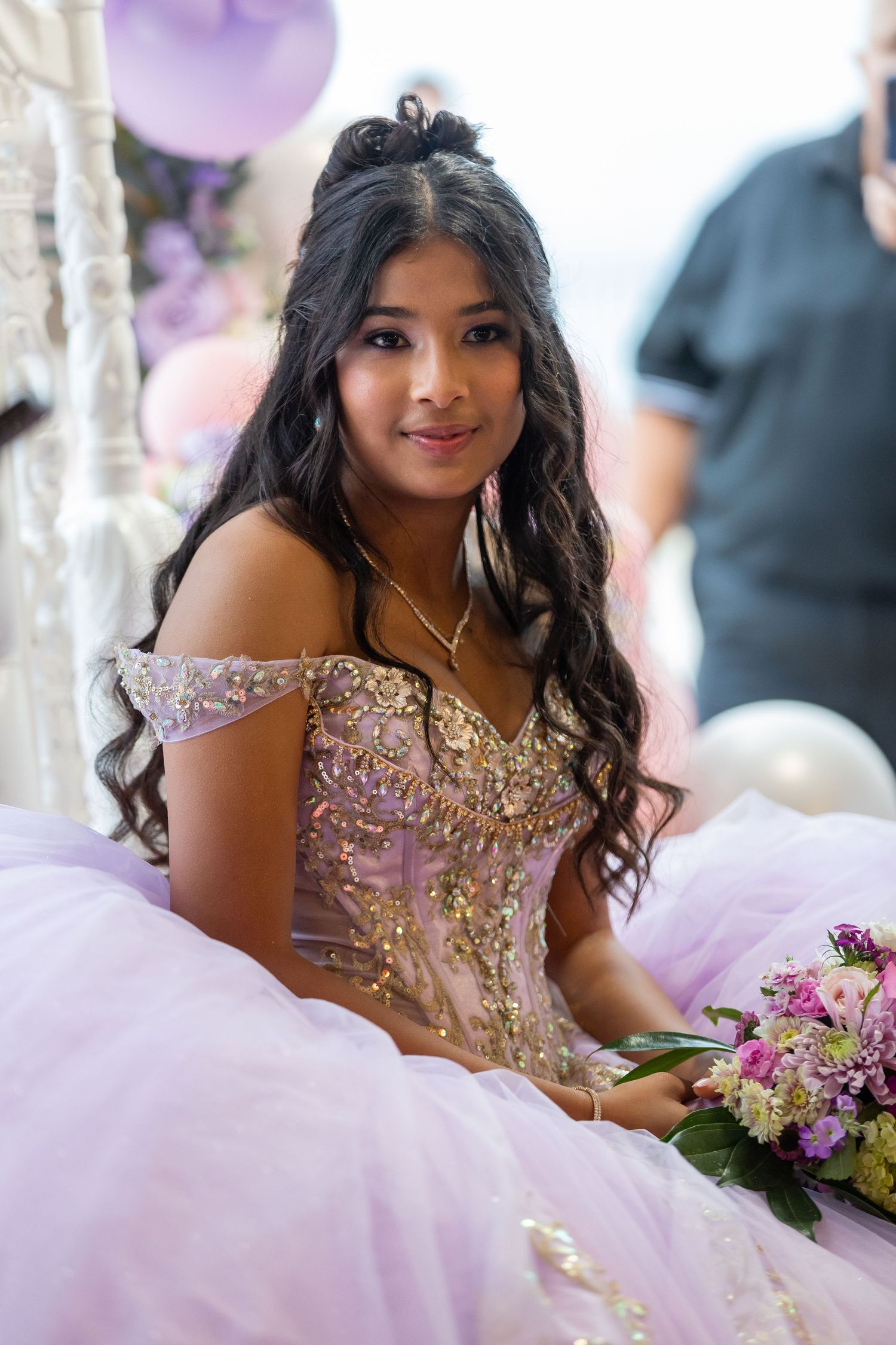 A quinceañera celebration features a young person in a sparkling beaded gown posing with purple floral decorations.