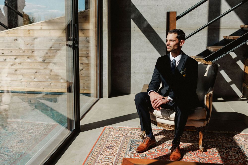 groom sits on a leather chair near a window with sunlight streaming in herdade do moinho novo
