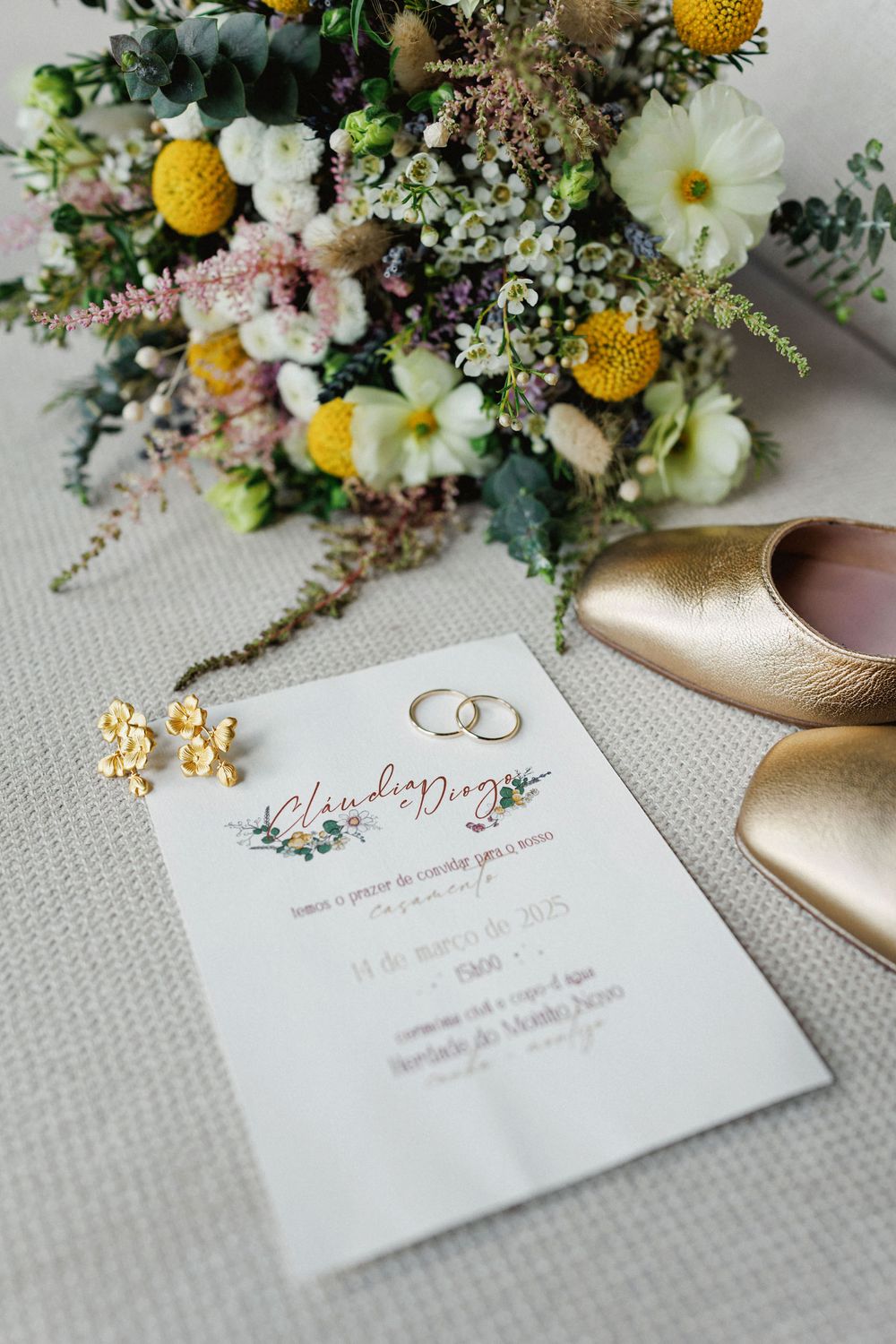 Wedding invitation with floral design lies beside gold shoes and yellow and white wildflower bouquet on textured fabric.