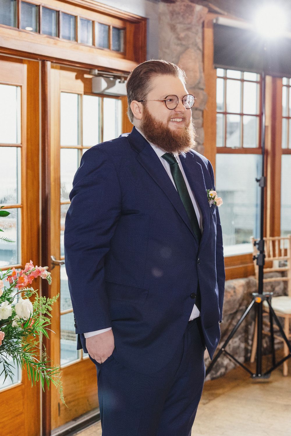Groom in navy suit and glasses poses in rustic venue with wooden windows and natural lighting.